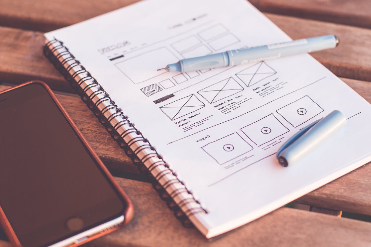services-04 Close-up of a notebook with wireframe sketches and a smartphone on a wooden desk.